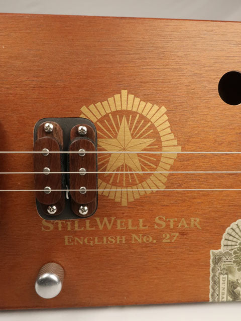 Close-up of a wooden cigar box guitar instrument with 'Stillwell Star' branding and 'English No. 27' text.