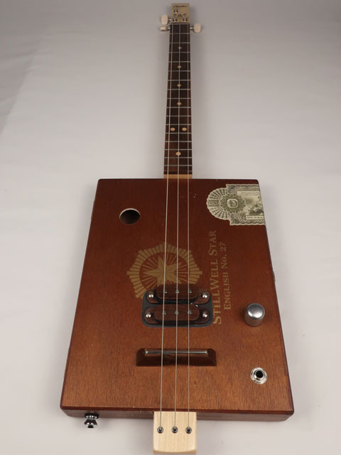 Brown wooden cigar box guitar with a visible brand logo on a white background. This cigar box guitar is handmade by Mike Snowden.