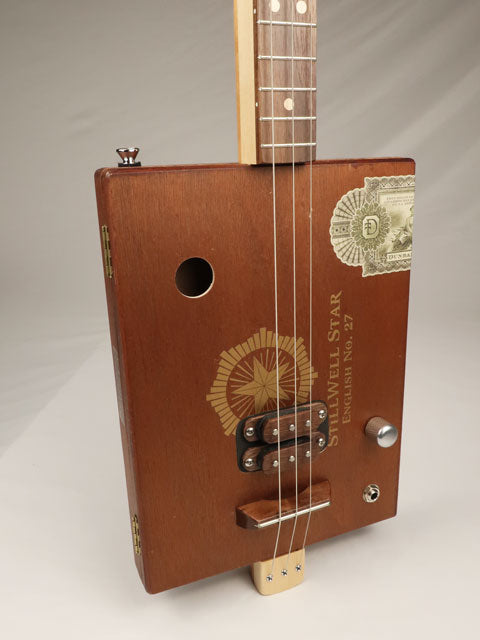 Brown wooden cigar box guitar with visible brand logo on a gray background. This Stillwell Star cigar box guitar is made by Mike Snowden.