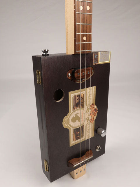 Front tilted view of a 3 string cigar box guitar made with a Pura Raza cigar box. This cigar box guitar is made with walnut and maple with a single coil pickup by Mike Snowden of Snowden Guitars.