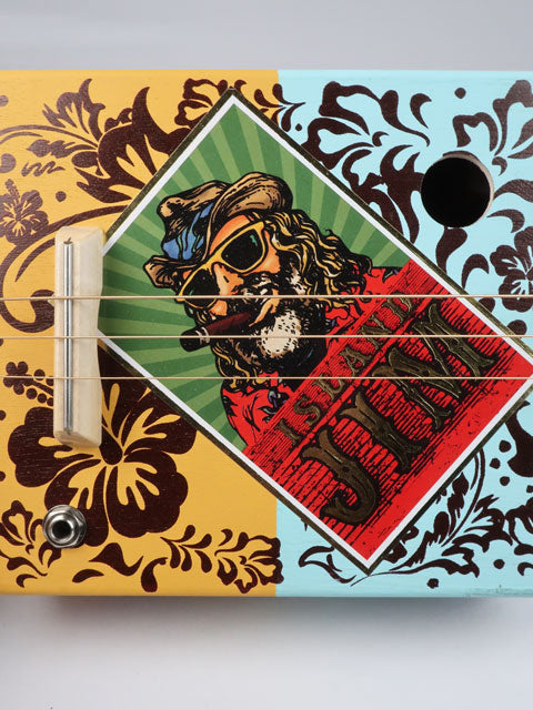Close up view of a cigar box guitar handmade by Mike Snowden. This is the front view with an Island Jim logo on the front with a maple bridge and strings. 