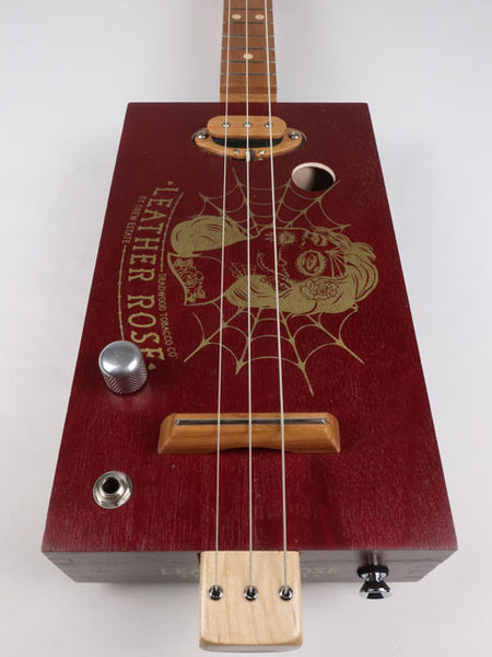 Front view of a Leather Rose 3 string cigar box guitar. Red painted cigar box with gold Leather Rose graphic on the front. This cigar box guitar is handmade by Mike Snowden of Snowden Guitars.