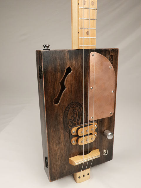 Wooden three string cigar box guitar with a f hole and copper pick guard on a gray background tilted view. Made by Mike Snowden with a Padron cigar box.